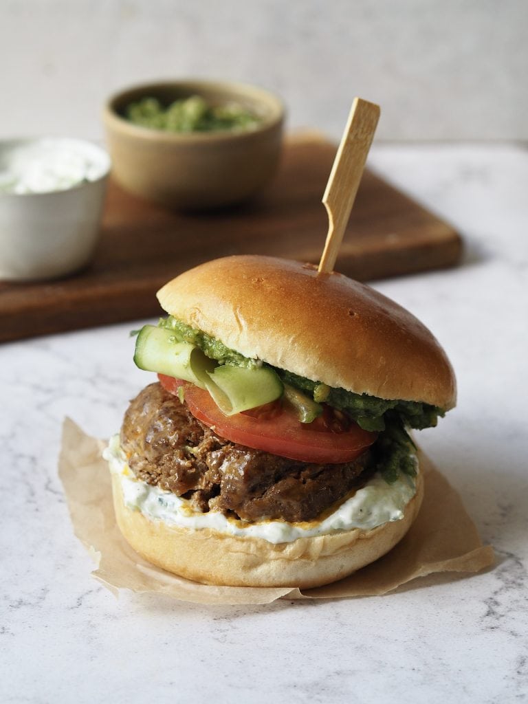 Lamb burger with tomato, cucumber ribbons, tzatziki and pesto on a white marble countertop.