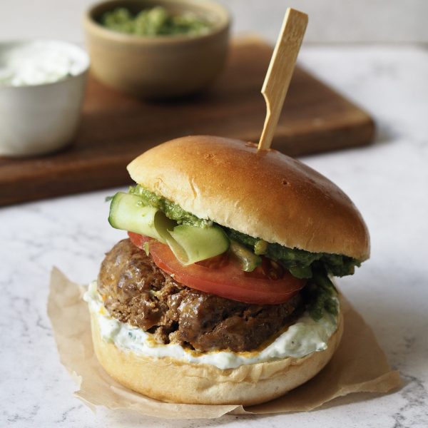 Lamb burger with tomato, cucumber ribbons, tzatziki and pesto on a white marble countertop.