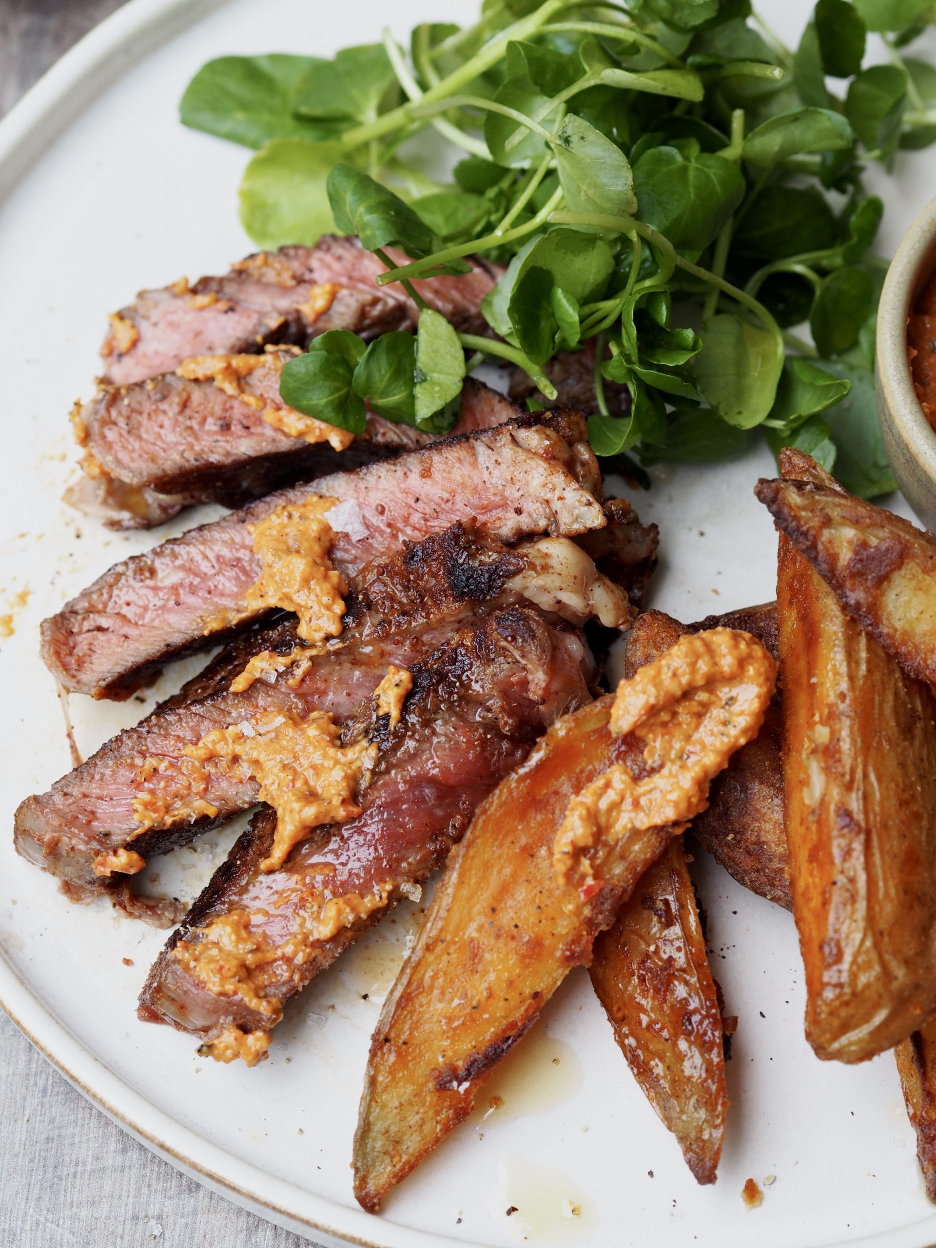 Close up of sliced steak, potato wedges and watercress on a white plate smeared with romesco sauce.