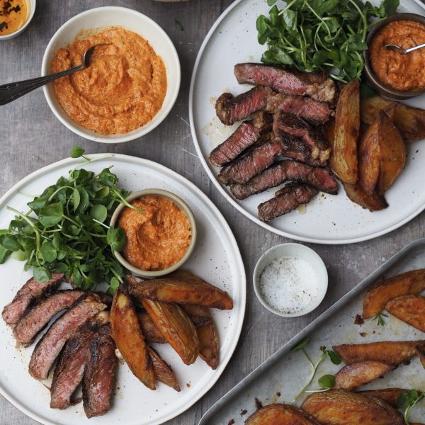 Plates of sliced rib eye steak with watercress, potato wedges and romesco sauce.