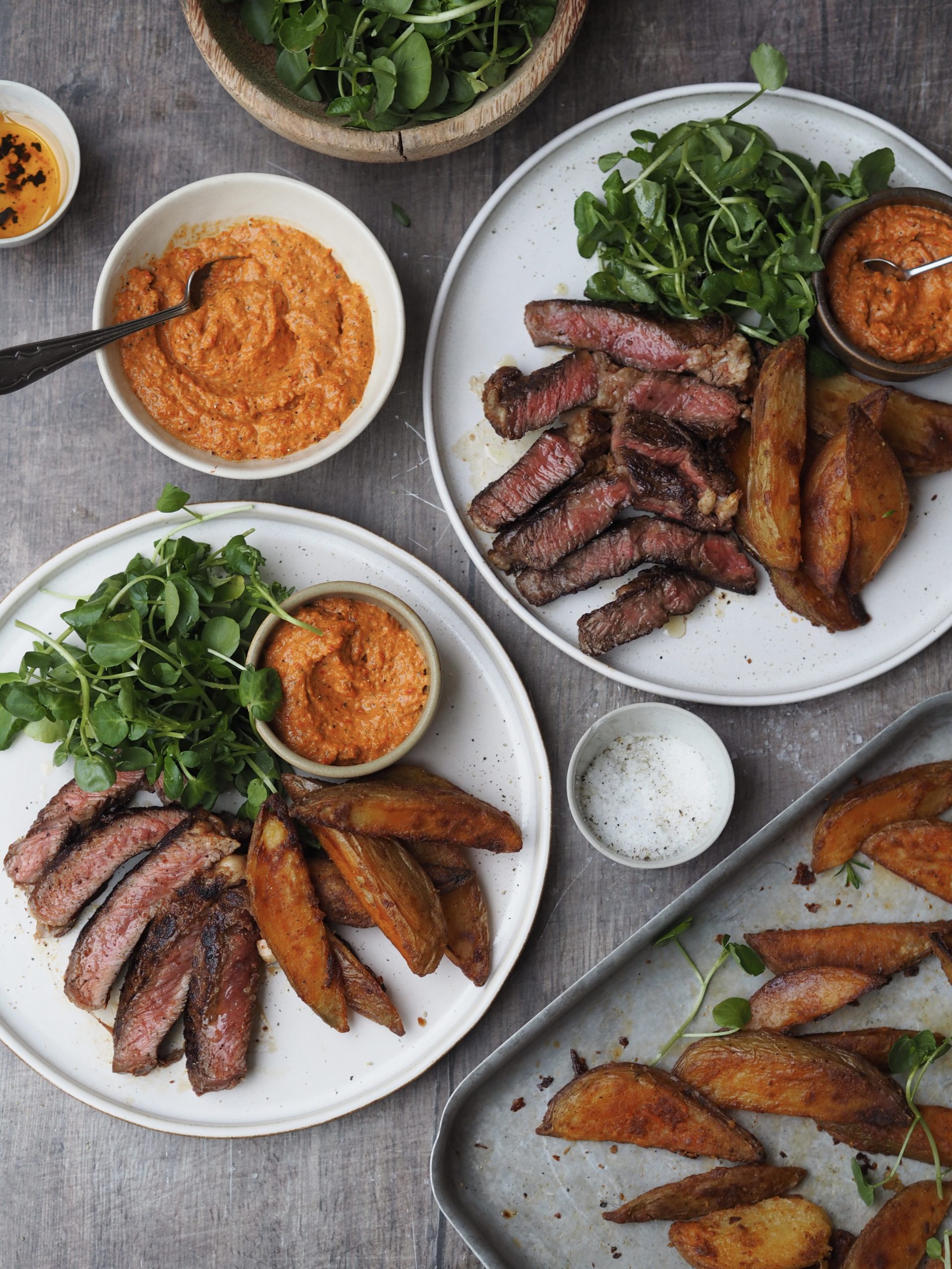 Plates of sliced rib eye steak with watercress, potato wedges and romesco sauce.