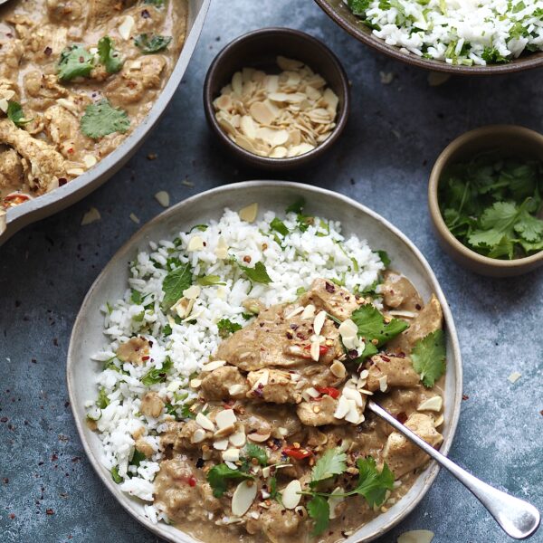 Plate of almond butter chicken curry and rice surrounded by serving dishes of rice, curry and toppings.