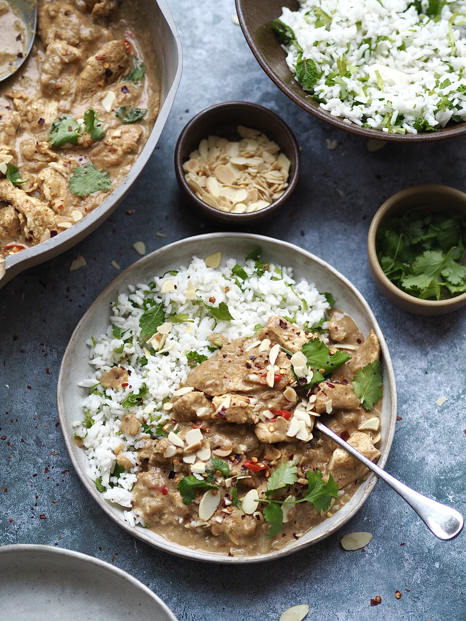 Plate of almond butter chicken curry and rice surrounded by serving dishes of rice, curry and toppings. 