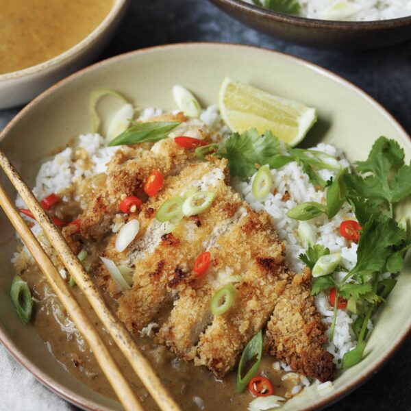 Chicken katsu cutlet in slices served over a bowl of rice with spring onions, chilli slices, lime wedges and coriander as a garnish.