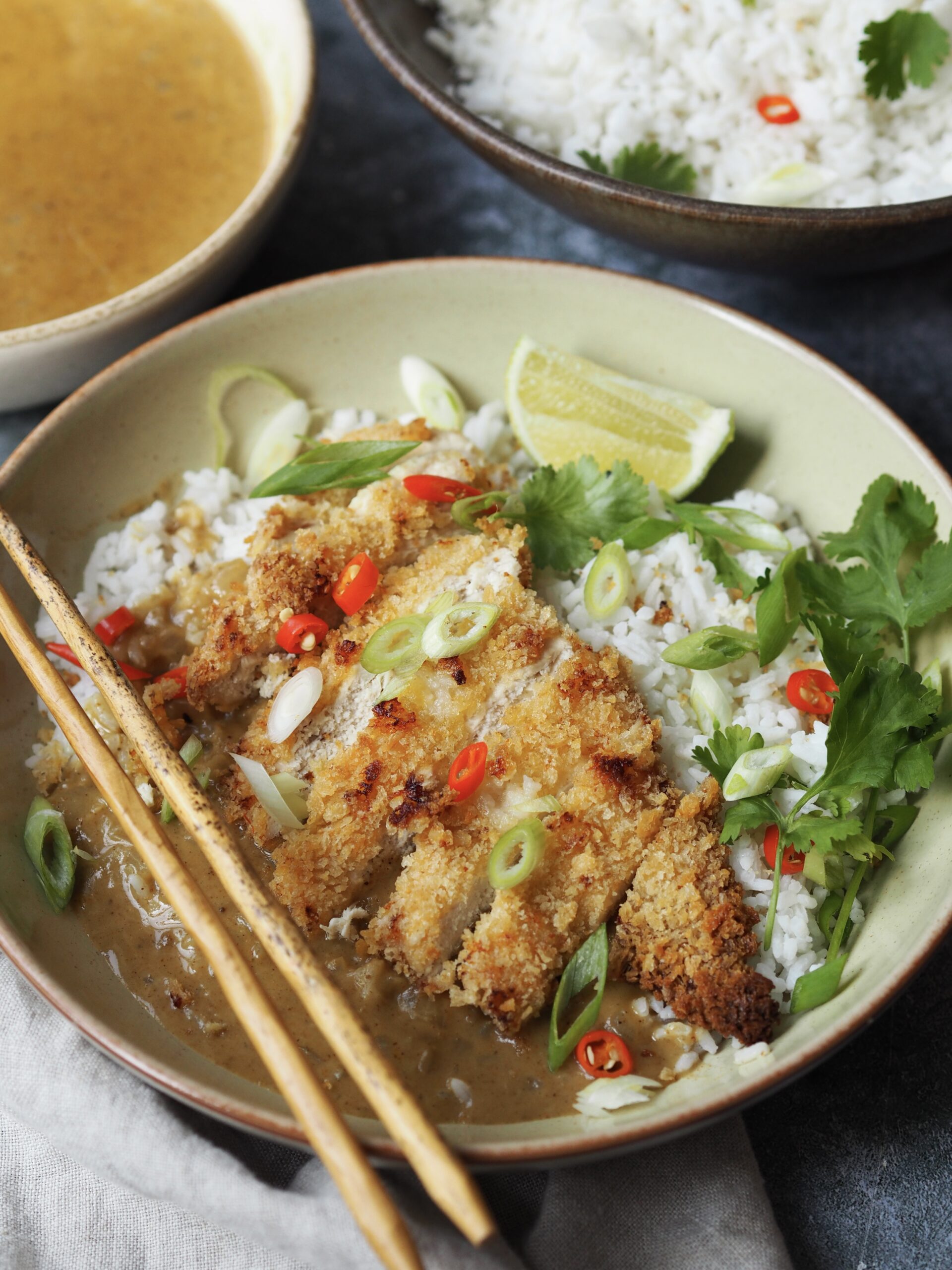 Chicken katsu cutlet in slices served over a bowl of rice with spring onions, chilli slices, lime wedges and coriander as a garnish. 