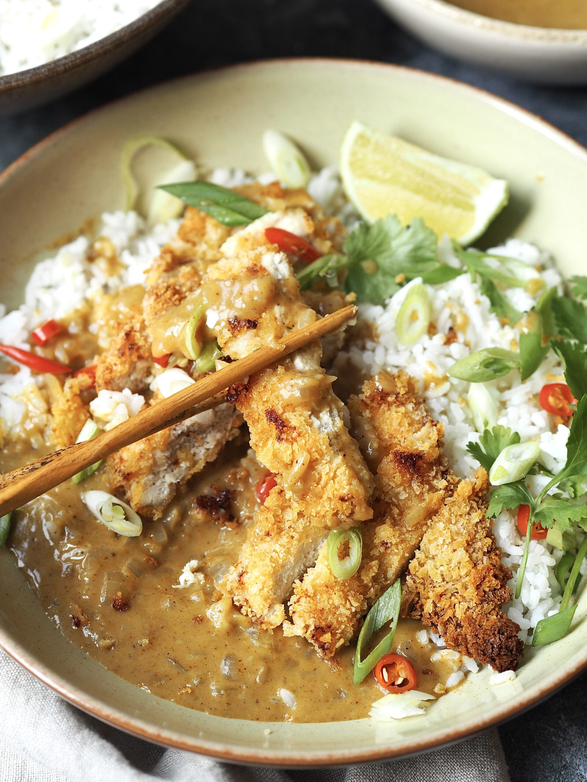 Wooden chopsticks picking up a piece of chicken katsu from a curry rice bowl.