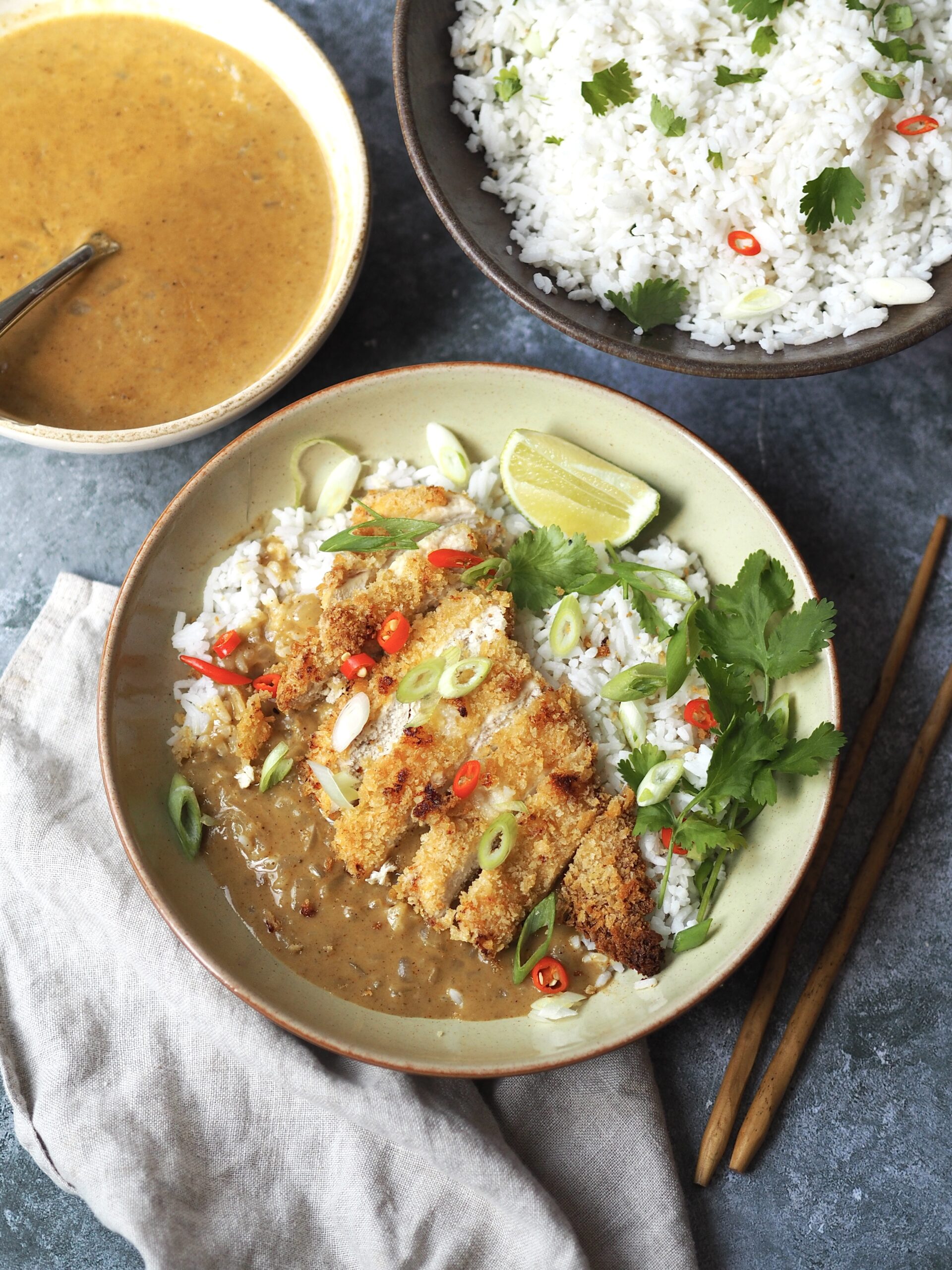 A chicken katsu curry bowl next to serving bowls of herbed rice and Japanese coconut curry sauce. 