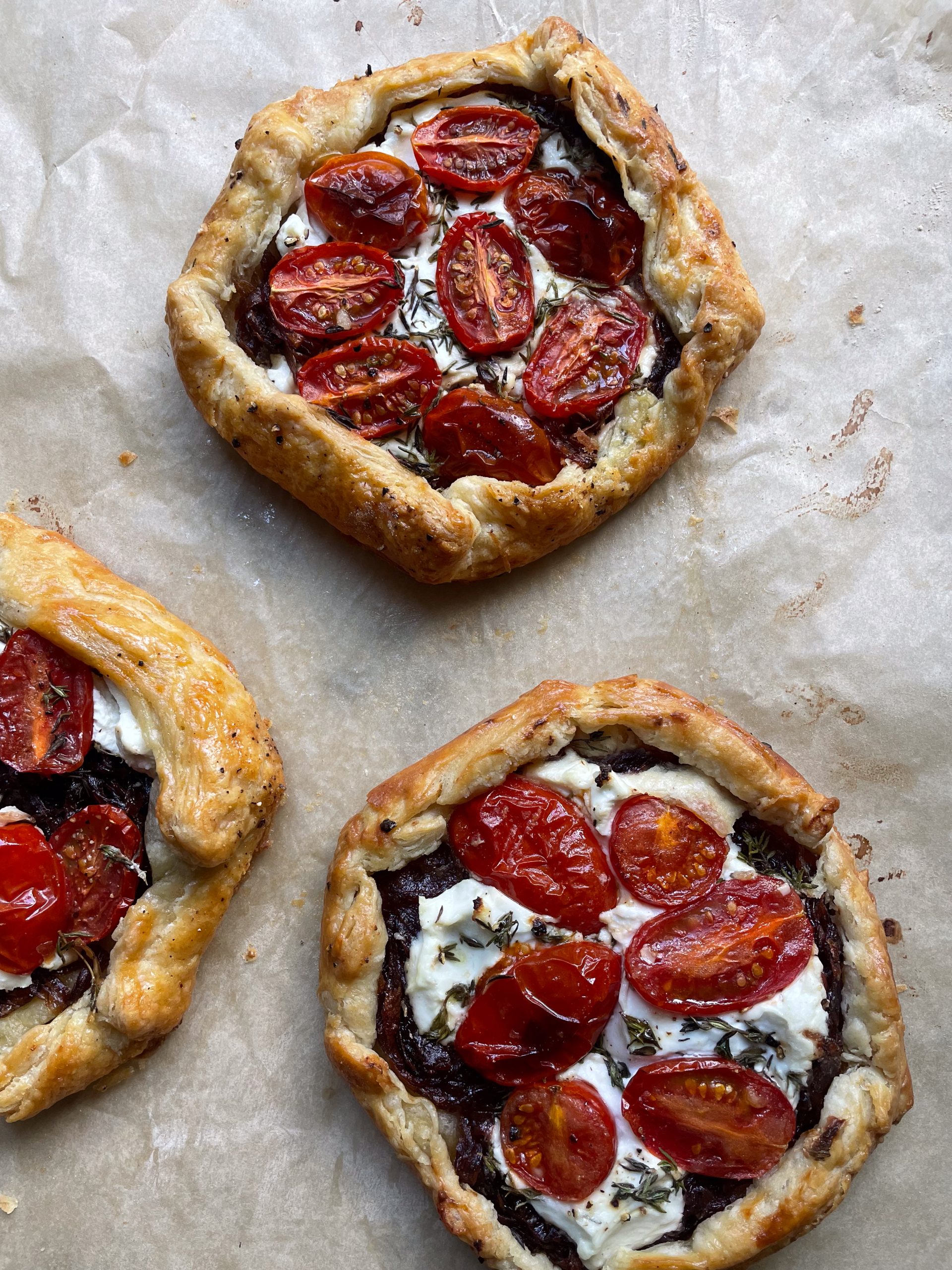 Three galettes with cherry tomatoes, goats cheese and red onions on a piece of baking parchment.