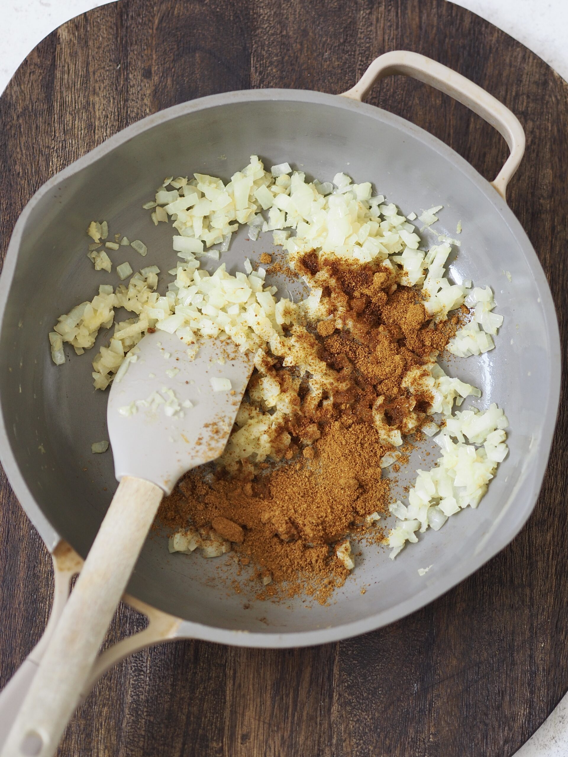 Curry powder being stirred into fried onions in a grey frying pan.