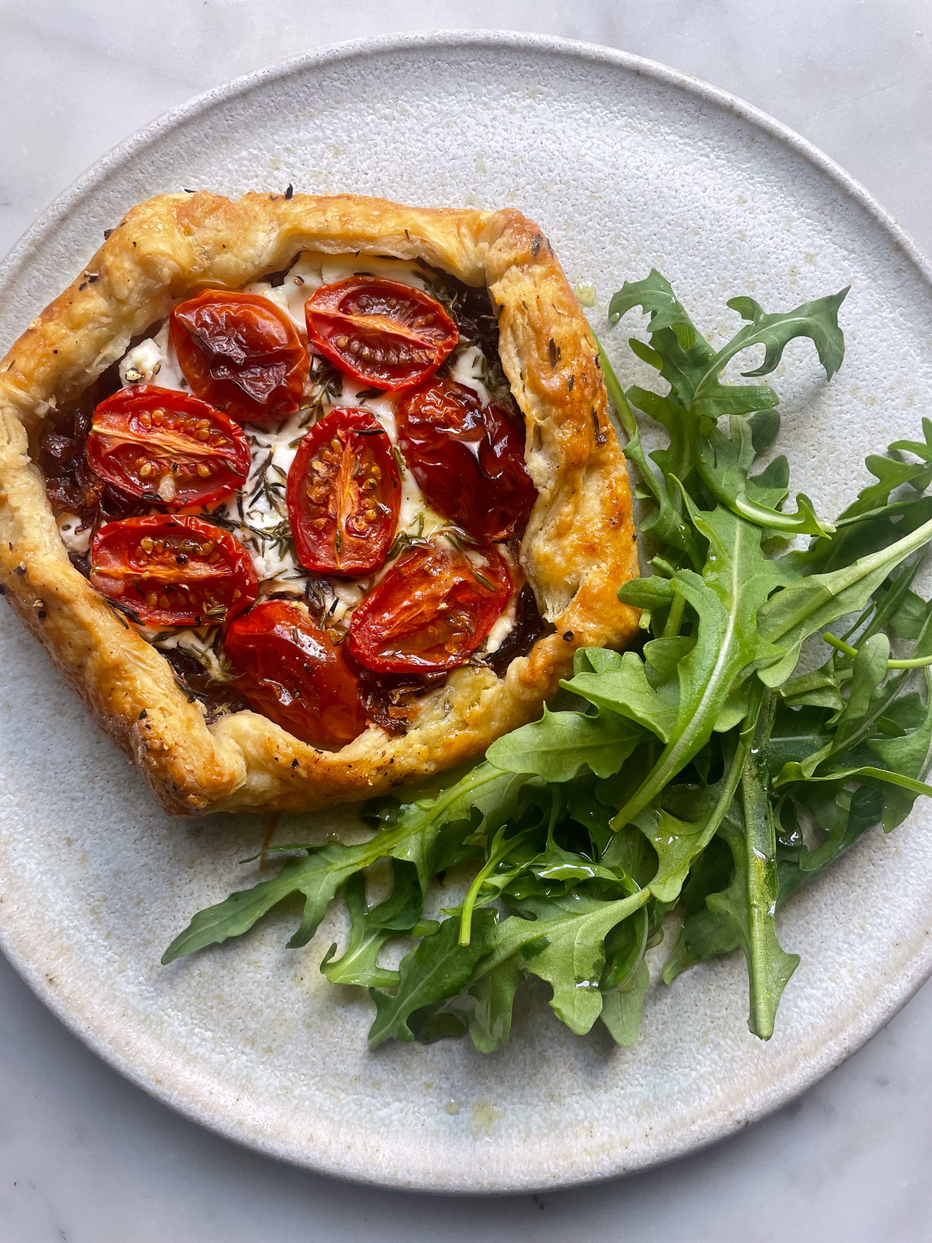 A red onion, goats cheese and cherry tomato galette on a grey plate with a side of rocket.