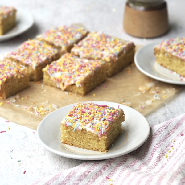 Slice of rainbow sprinkle iced sponge on a white plate with the rest of the cake in the background.