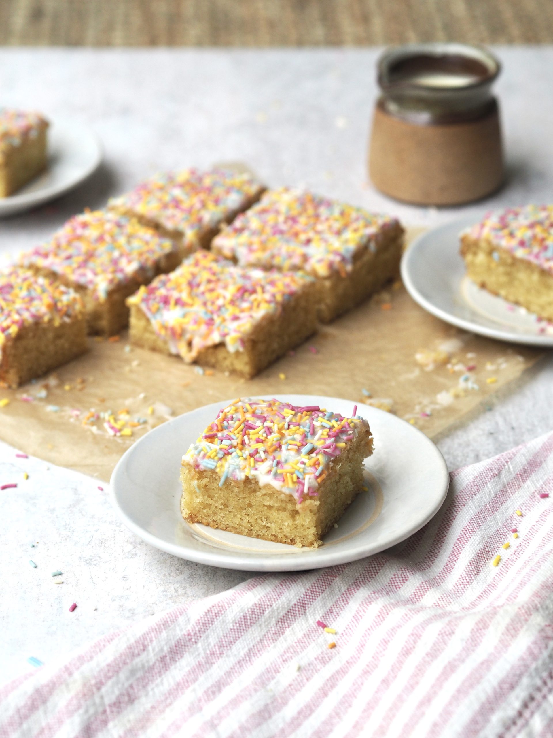 Slice of rainbow sprinkle iced sponge on a white plate with the rest of the cake in the background.