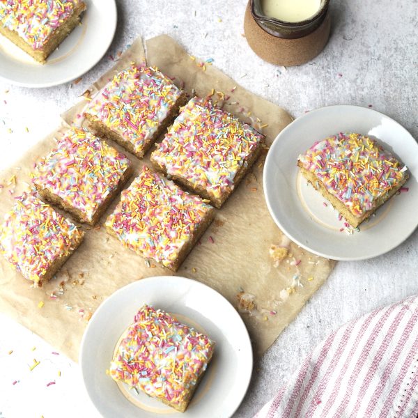 White plates of iced sprinkle sponge next to the cake cut into squares.