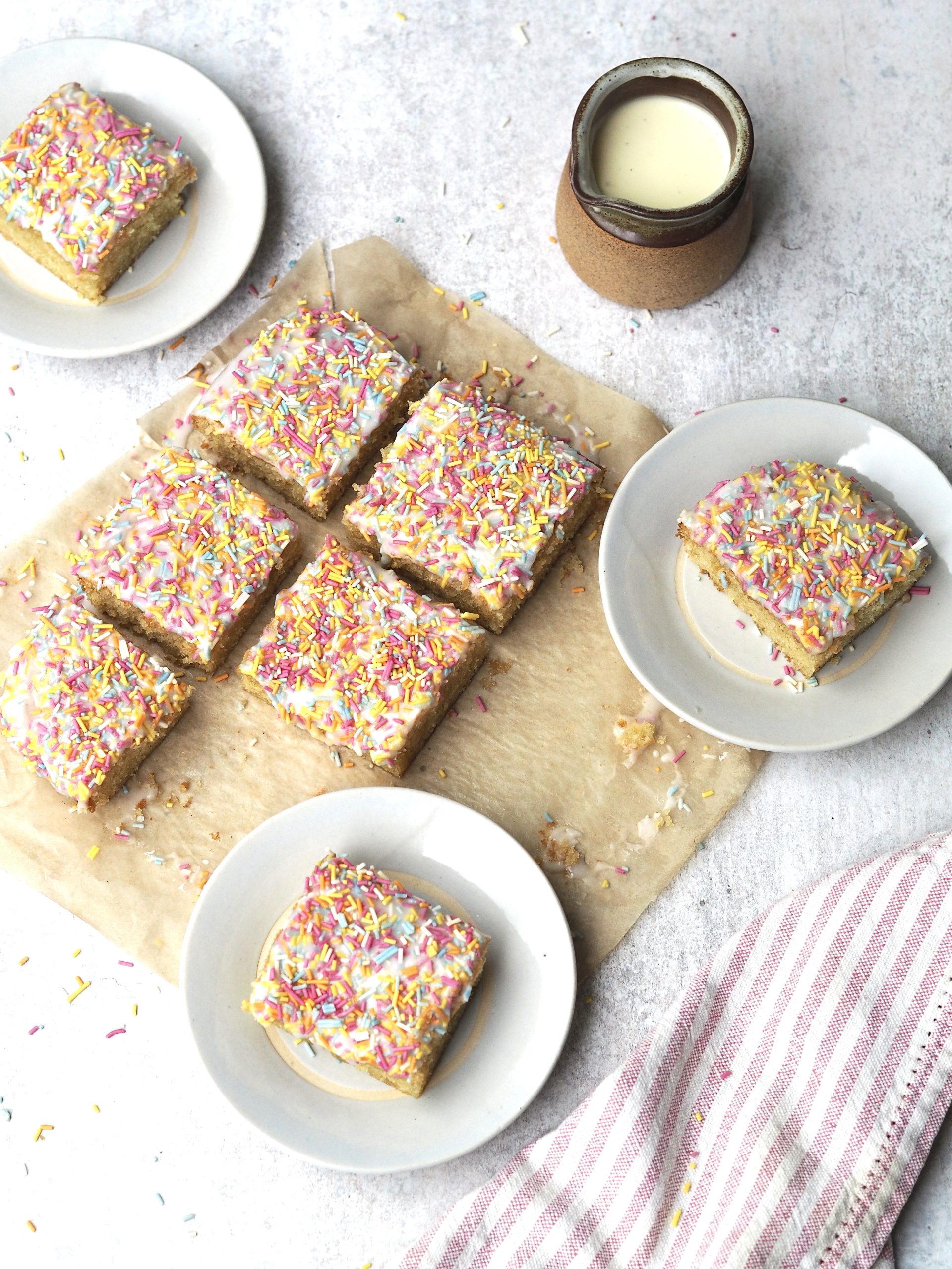 White plates of iced sprinkle sponge next to the cake cut into squares.