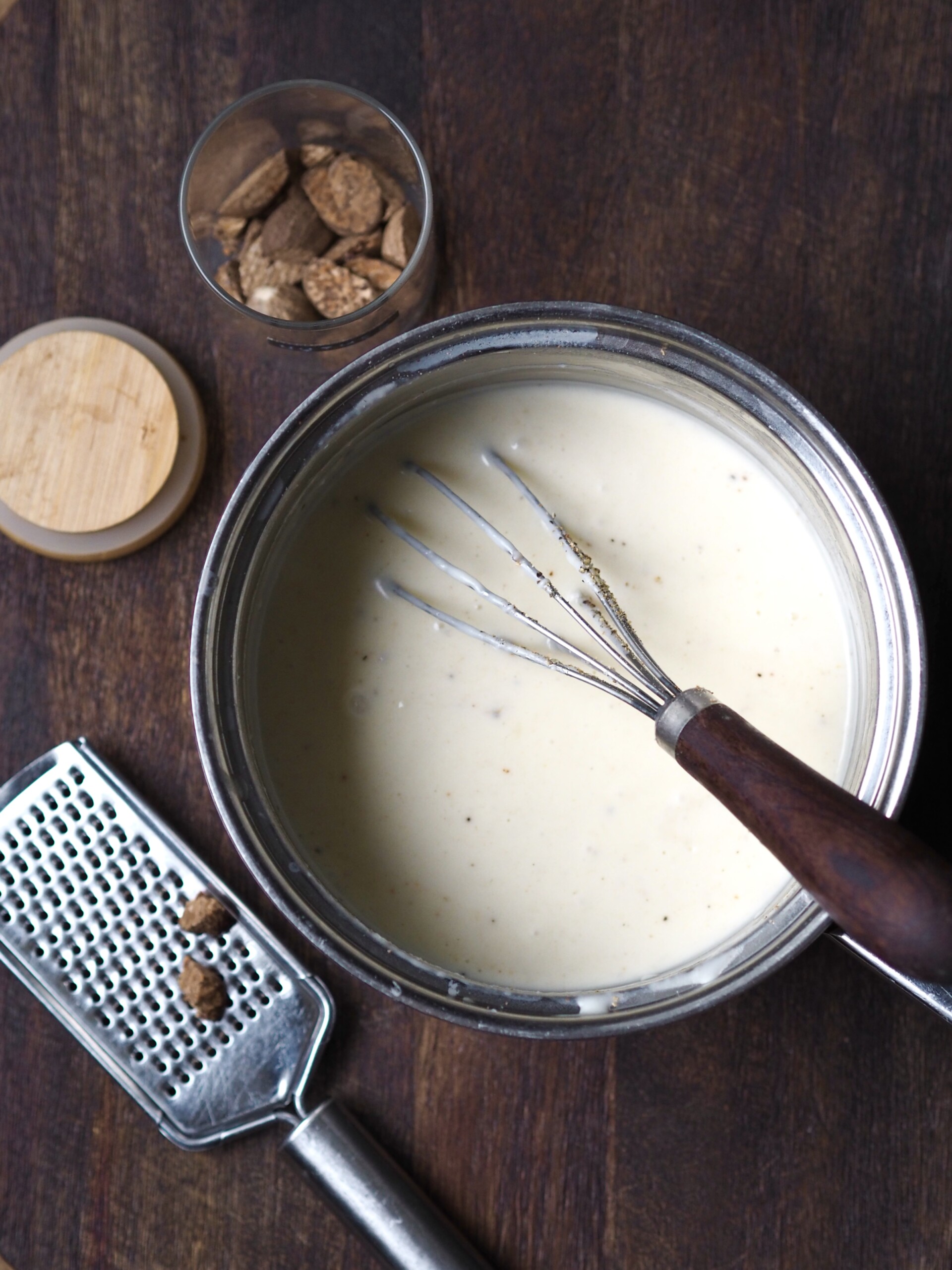 White sauce being whisked in a saucepan with the nutmeg and grater next to it.