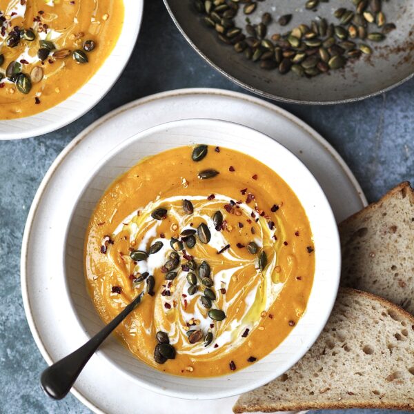 White bowl of butternut squash soup garnished with pumpkin seeds and chilli flakes with bread on the side.