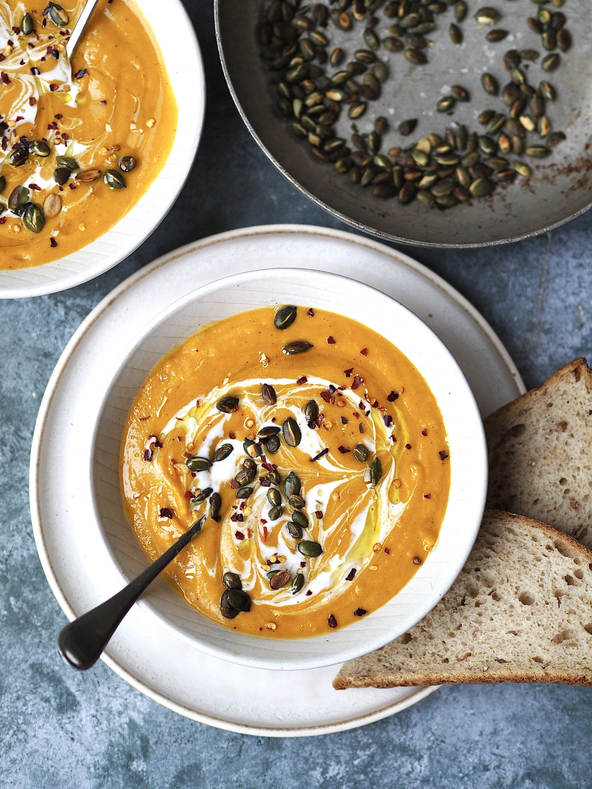 White bowl of butternut squash soup garnished with pumpkin seeds and chilli flakes with bread on the side.