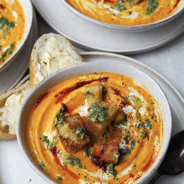 Bowl of carrot soup swirled with cream, harissa, and topped with pesto and croutons.