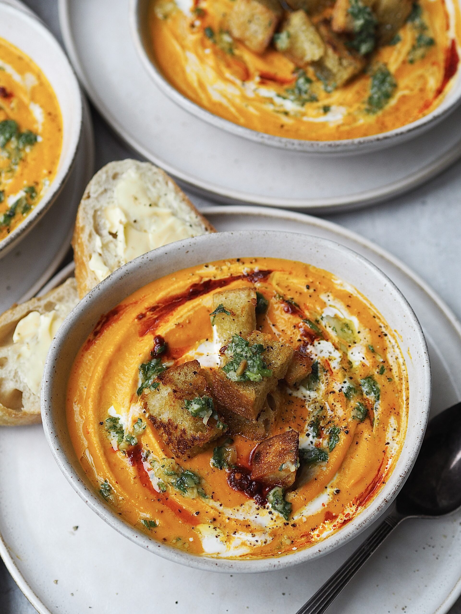 Bowl of carrot soup swirled with cream, harissa, and topped with pesto and croutons.