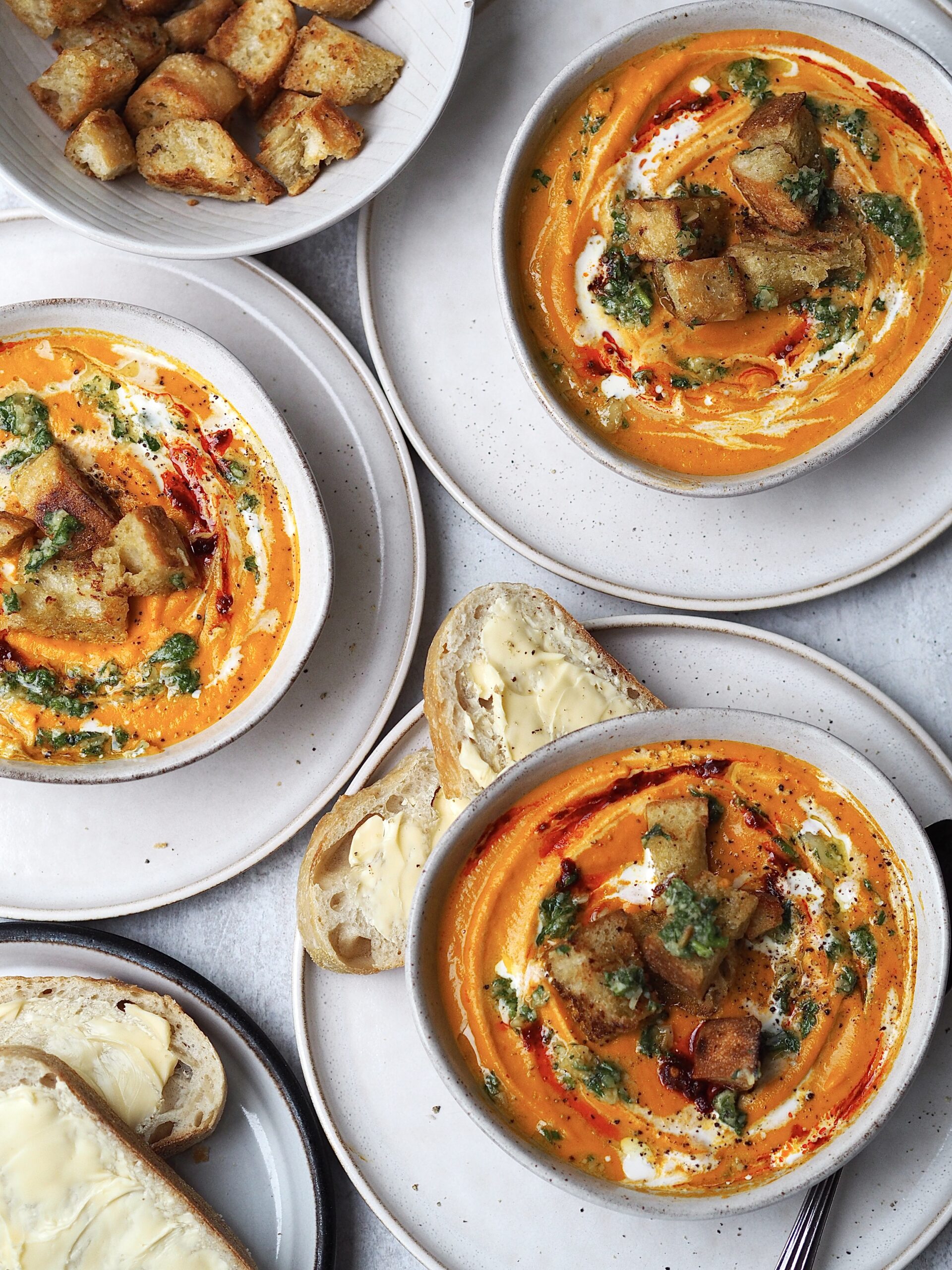 Grey bowls of roasted carrot and harissa soup topped with croutons and carrot top pesto.