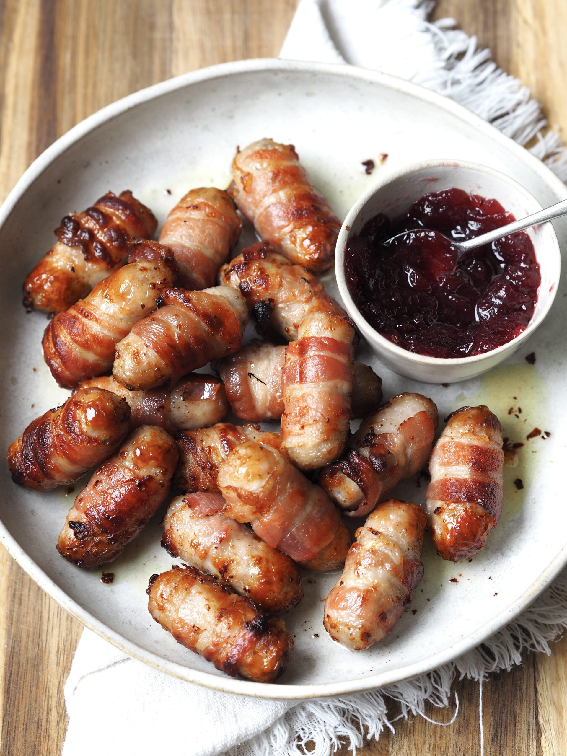 Close up of a white serving plate of pigs in blankets next to a bowl of cranberry sauce.