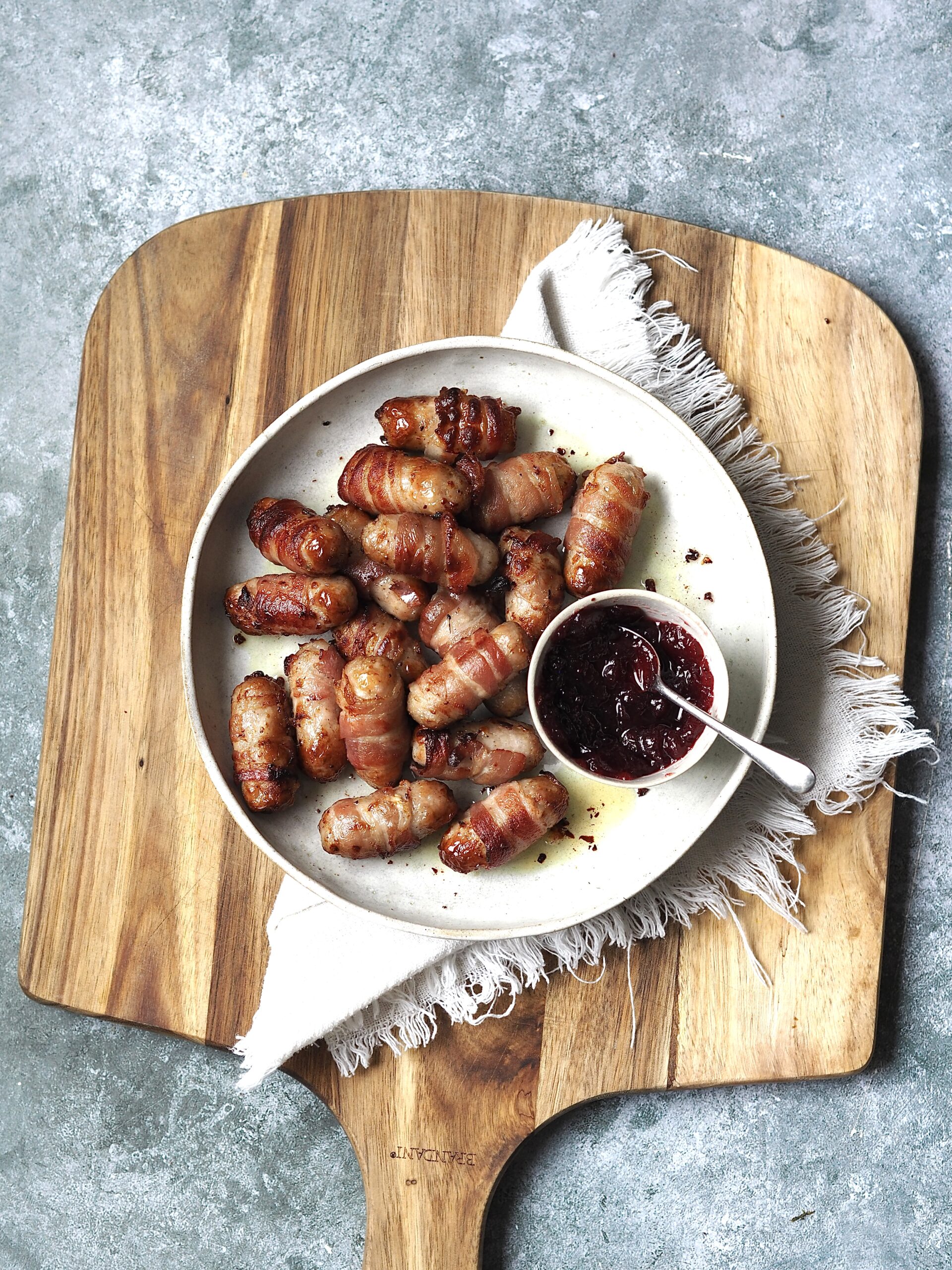 Wooden board with a plate of sausages wrapped in bacon with a dipping bowl of cranberry sauce.