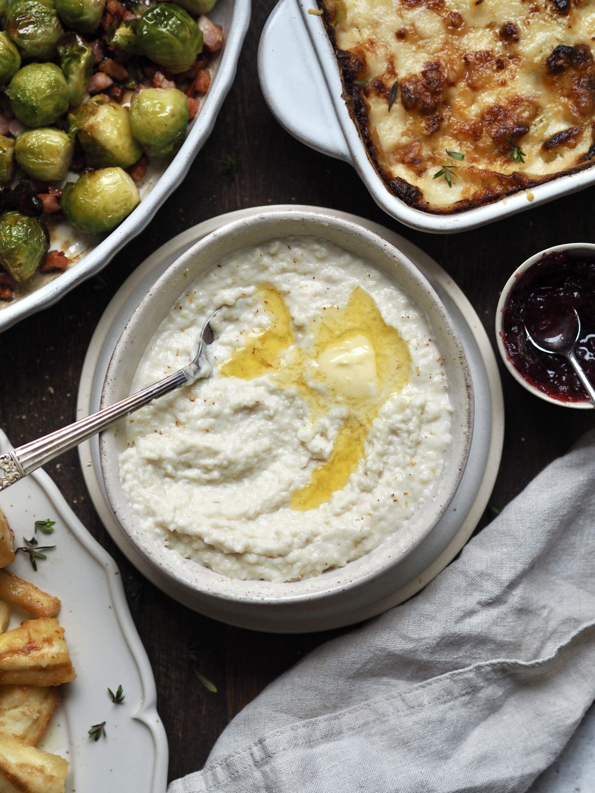 White bowl of bread sauce on a table with dishes of brussels sprouts, cheesy leeks, glazed parsnips and cranberry sauce.