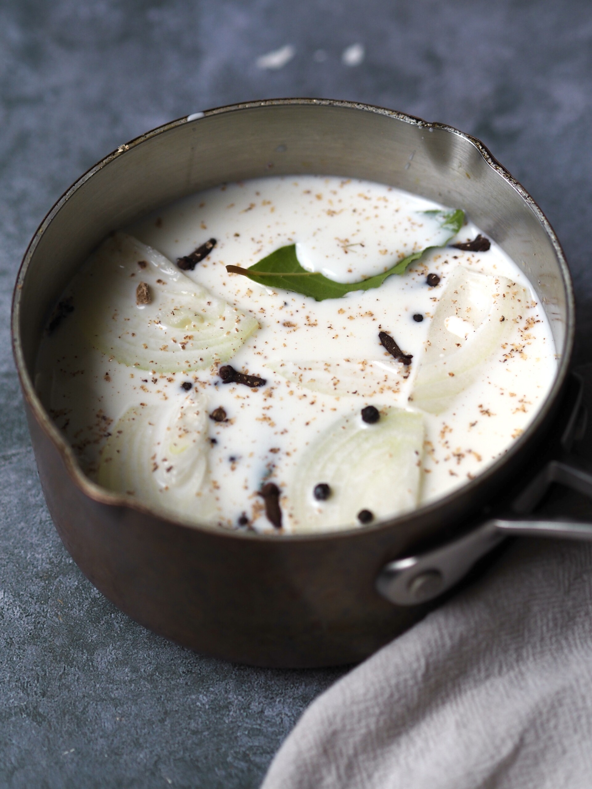 Saucepan of milk being infused with onion, peppercorns, nutmeg and a fresh bay leaf.