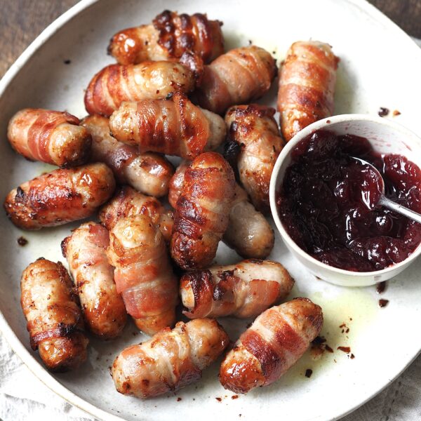 Sausages wrapped in bacon on a white serving plate next to a bowl of cranberry sauce.