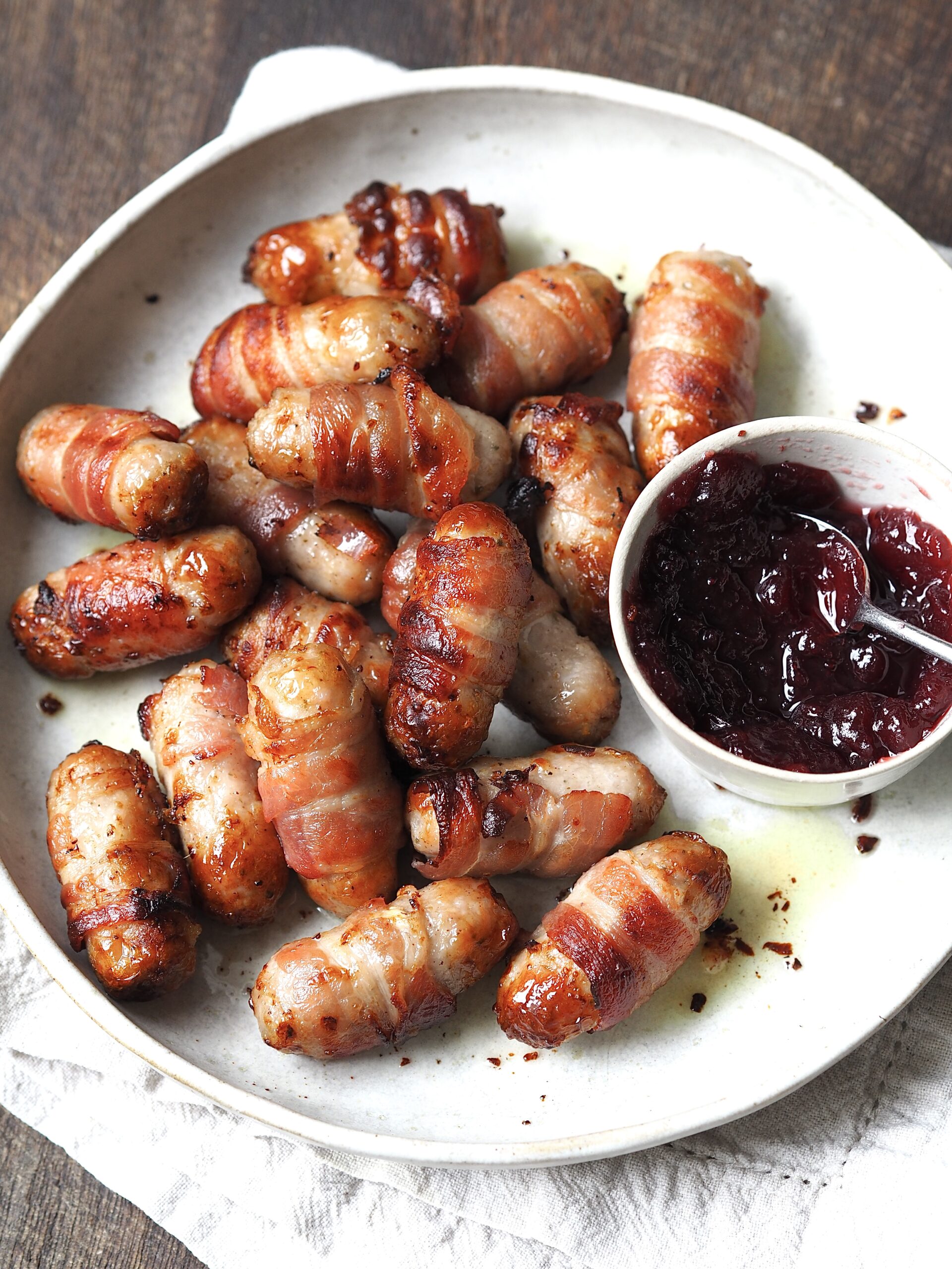 Sausages wrapped in bacon on a white serving plate next to a bowl of cranberry sauce.