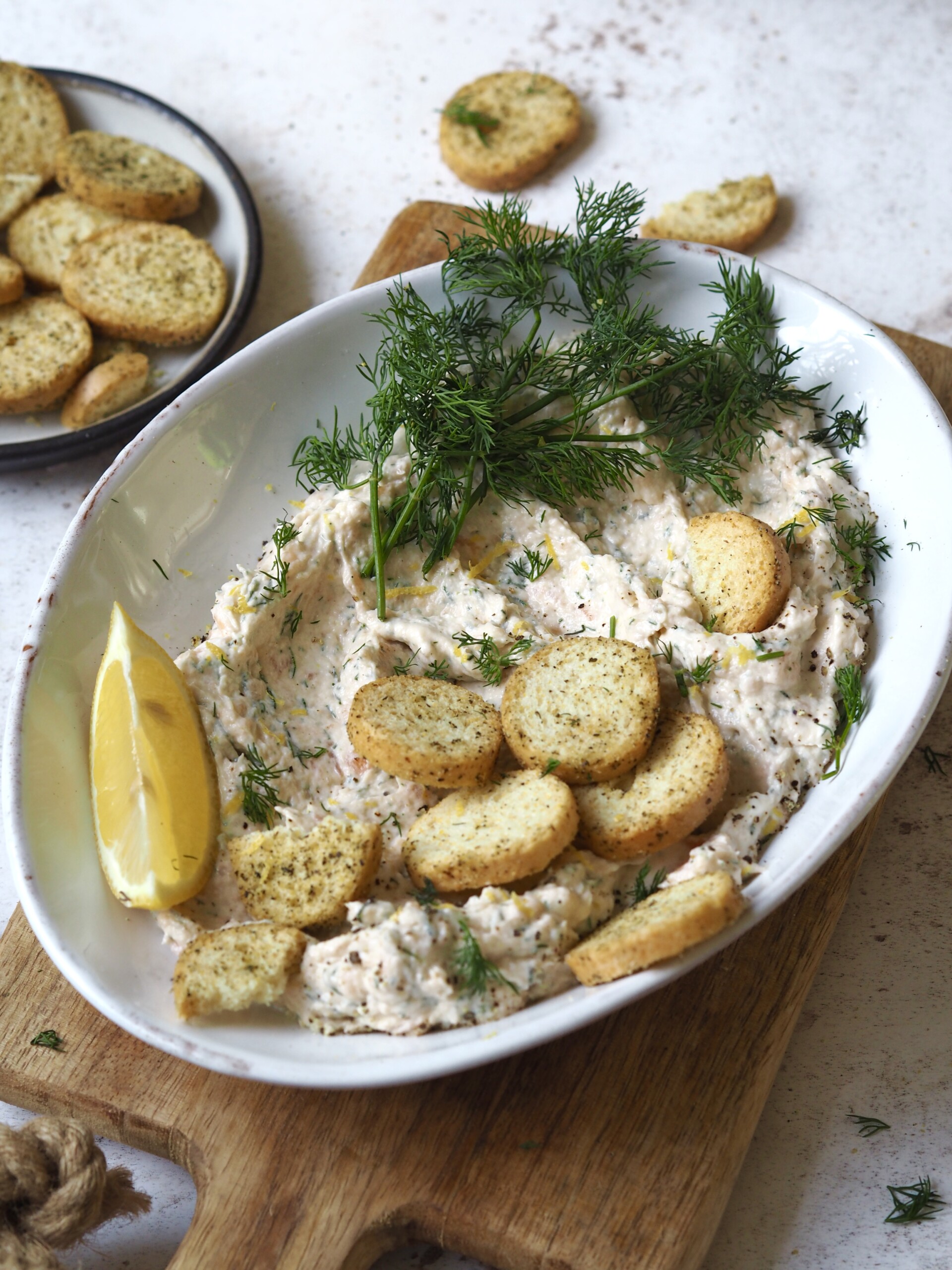 Oval bowl of smoked salmon pate with fresh dill, crostini and a lemon wedge on a wooden serving board.