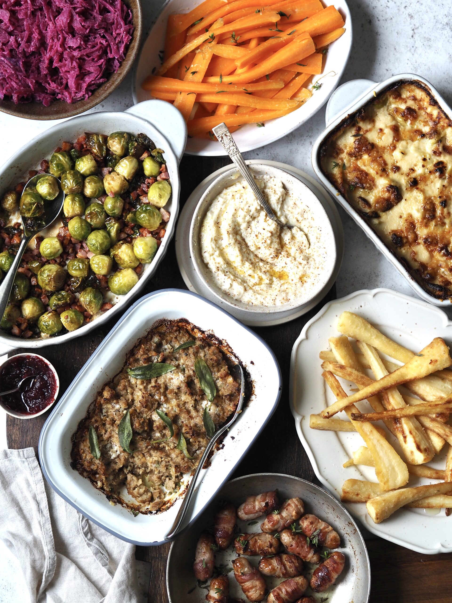 Christmas sides all gathered in serving dishes including red cabbage, glazed carrots, sprouts with bacon, bread sauce, cheesy leeks, baked stuffing, pigs in blankets and roasted parsnips.