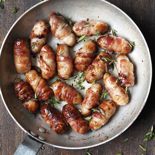 Metal saucepan on a wooden countertop with cooked pigs in blankets with a sticky fresh thyme glaze.