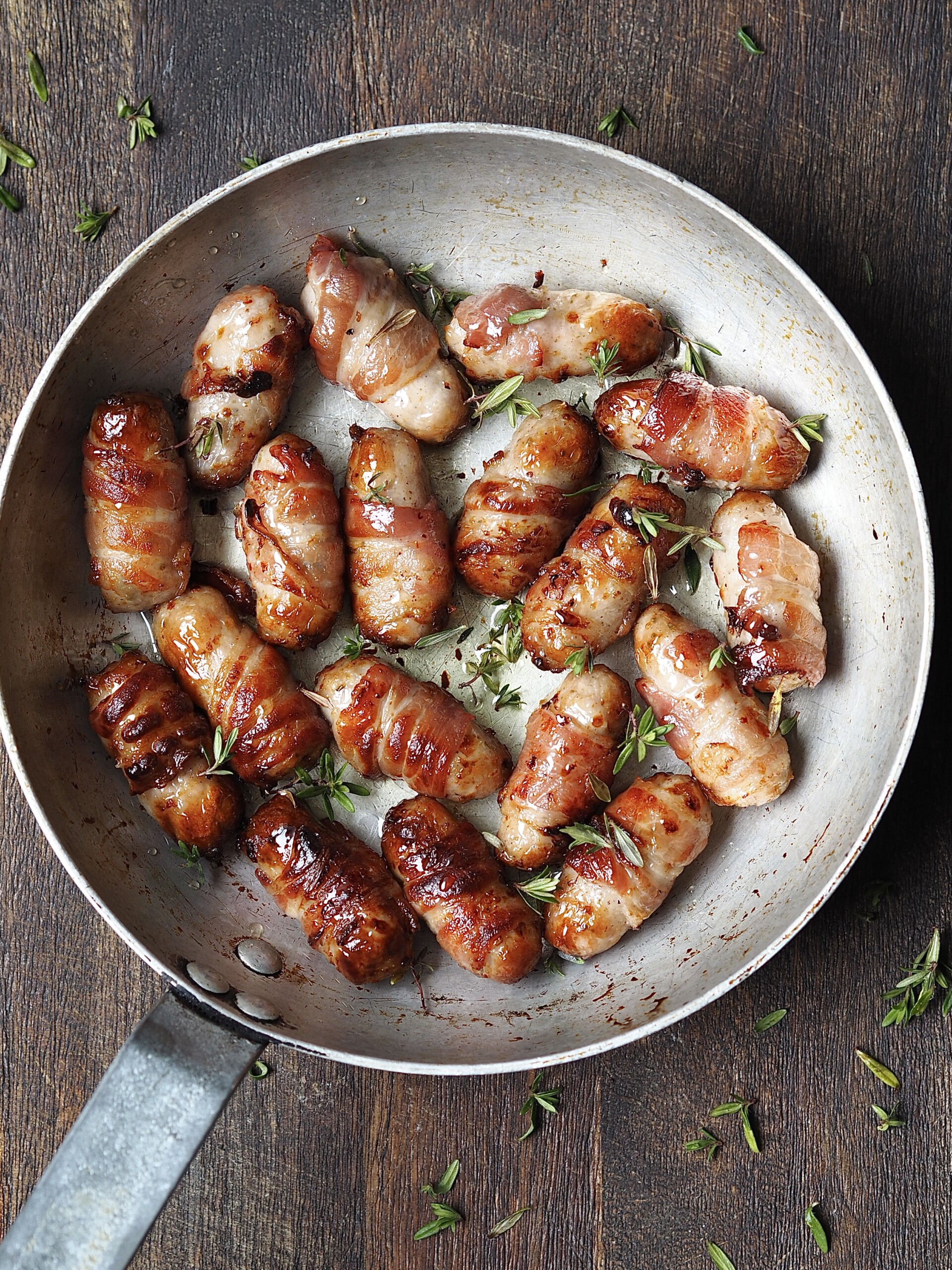 Metal saucepan on a wooden countertop with cooked pigs in blankets with a sticky fresh thyme glaze. 