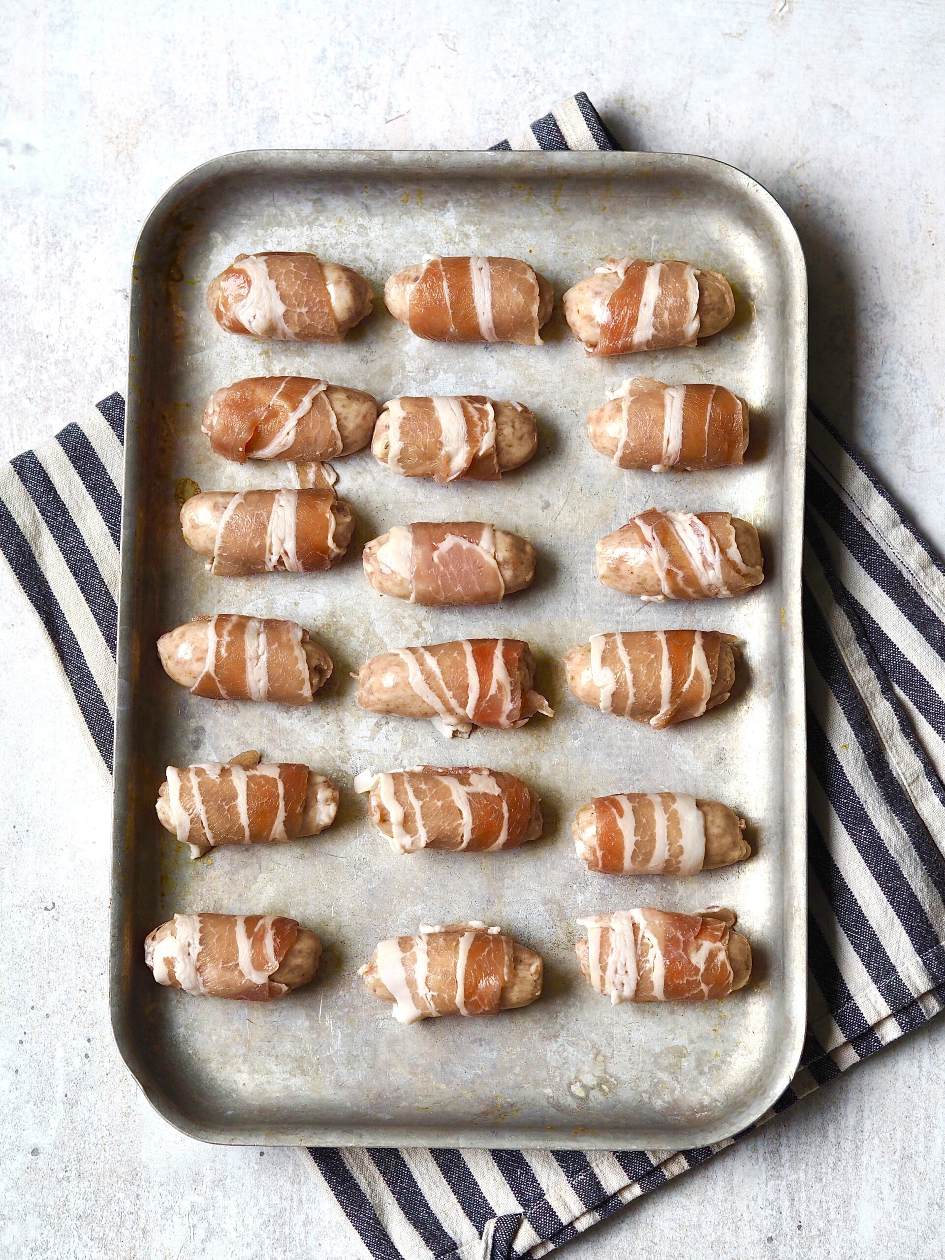 Metal roasting tray of uncooked pigs in blankets.