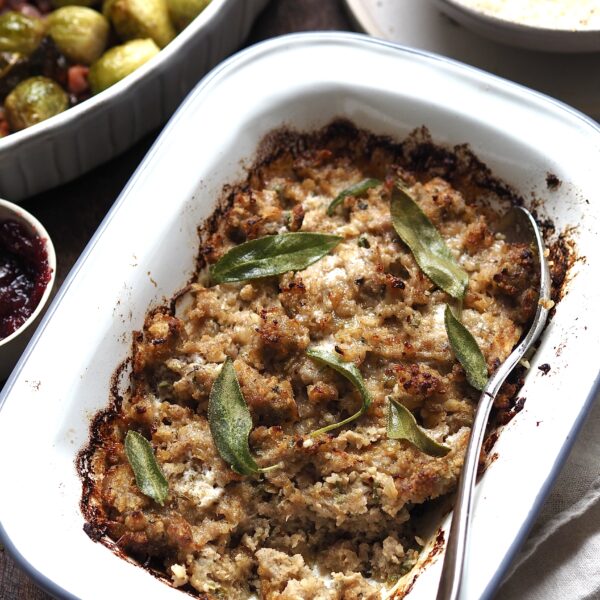 Sausage, Onion and Sage Stuffing in a white baking dish with cranberry sauce, brussels sprouts and bread sauce in the background.