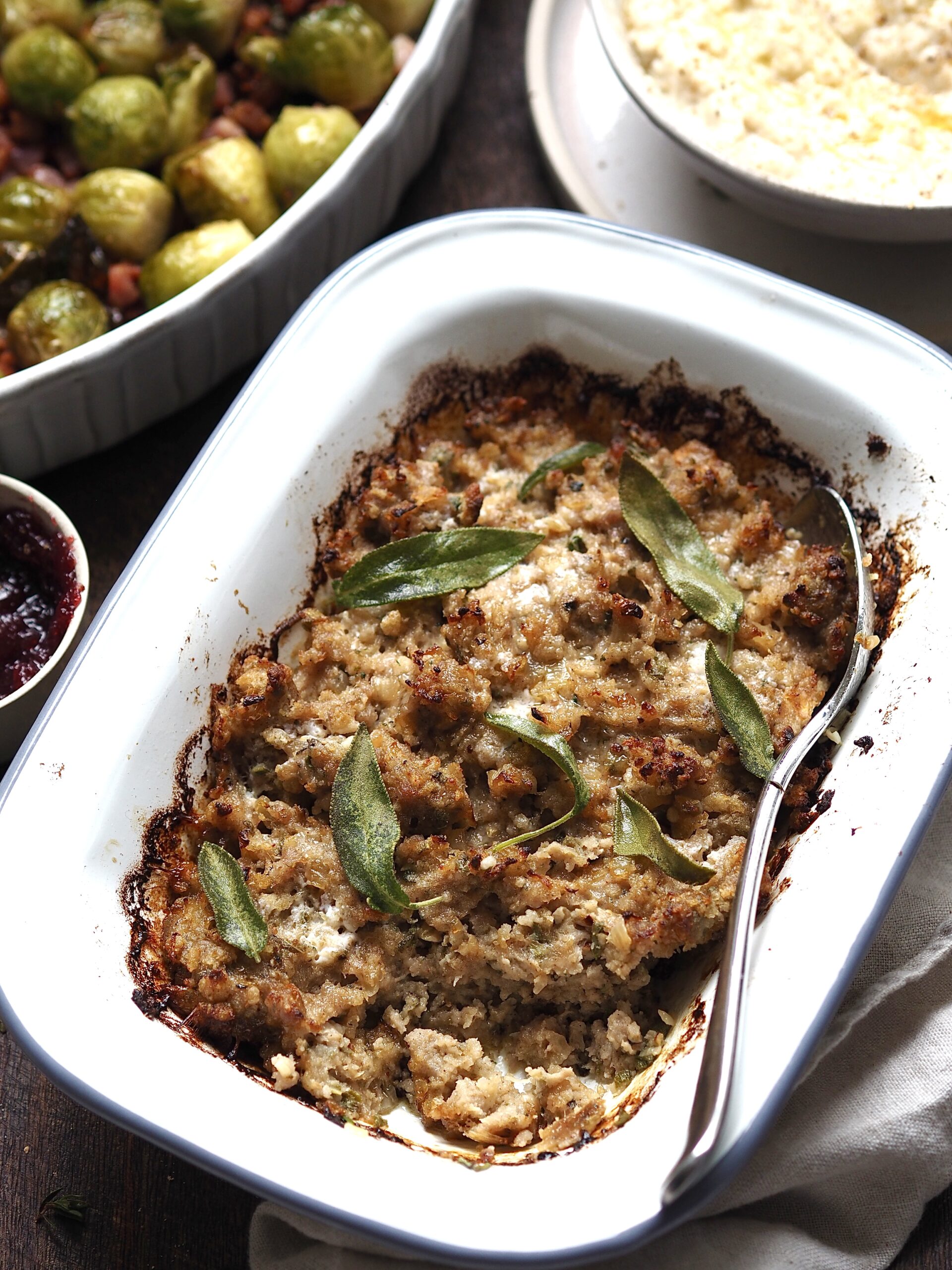 Sausage, Onion and Sage Stuffing in a white baking dish with cranberry sauce, brussels sprouts and bread sauce in the background.