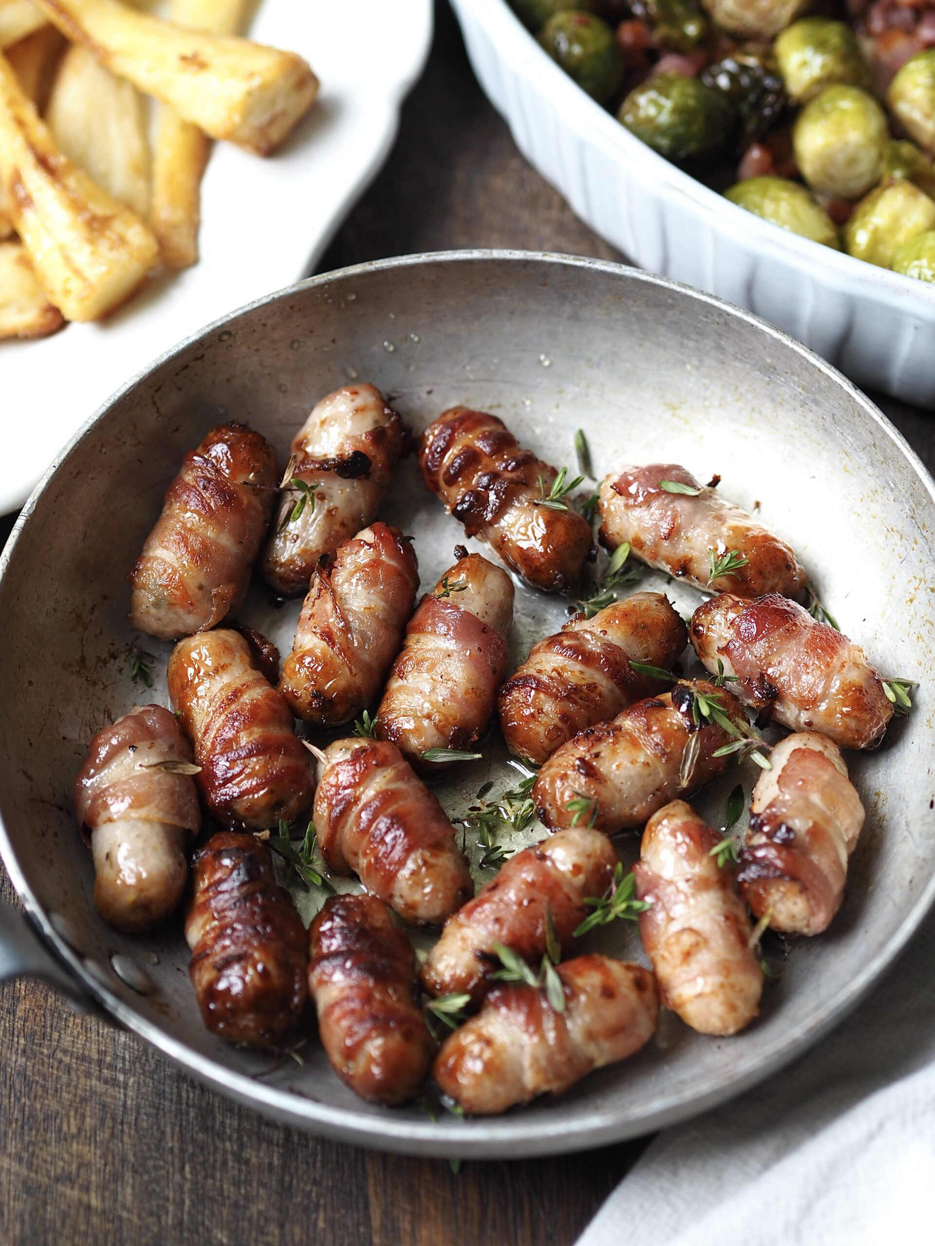 Pan of sticky pigs in blankets with dishes of parsnips and roasted sprouts in the background.