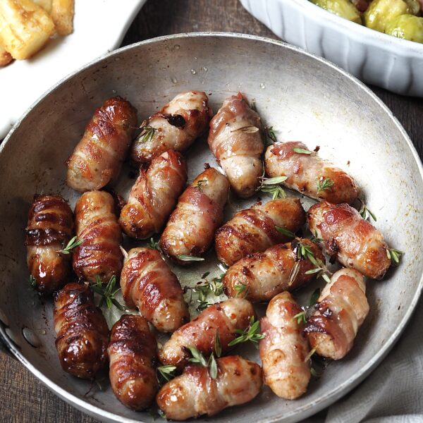 Frying pan of sticky pigs in blankets topped with fresh thyme next to white platters of bacon sprouts and roasted parsnips.