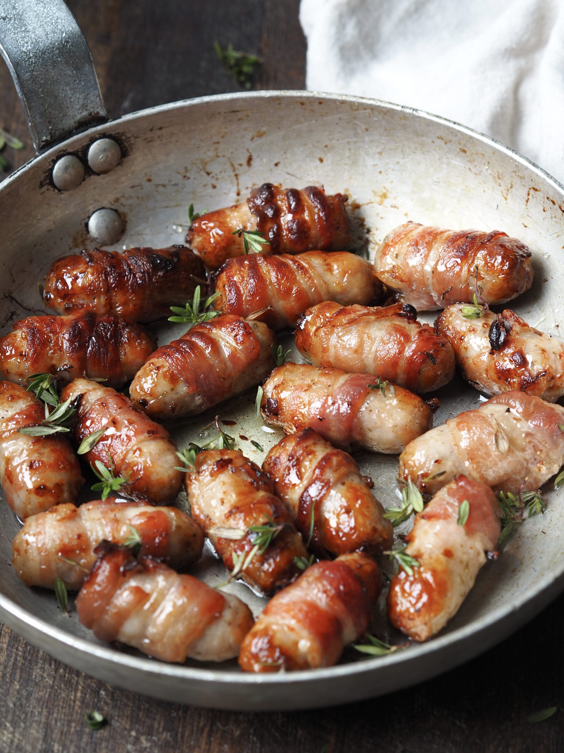 Side on view of a frying pan of sticky pigs in blankets with fresh rosemary.