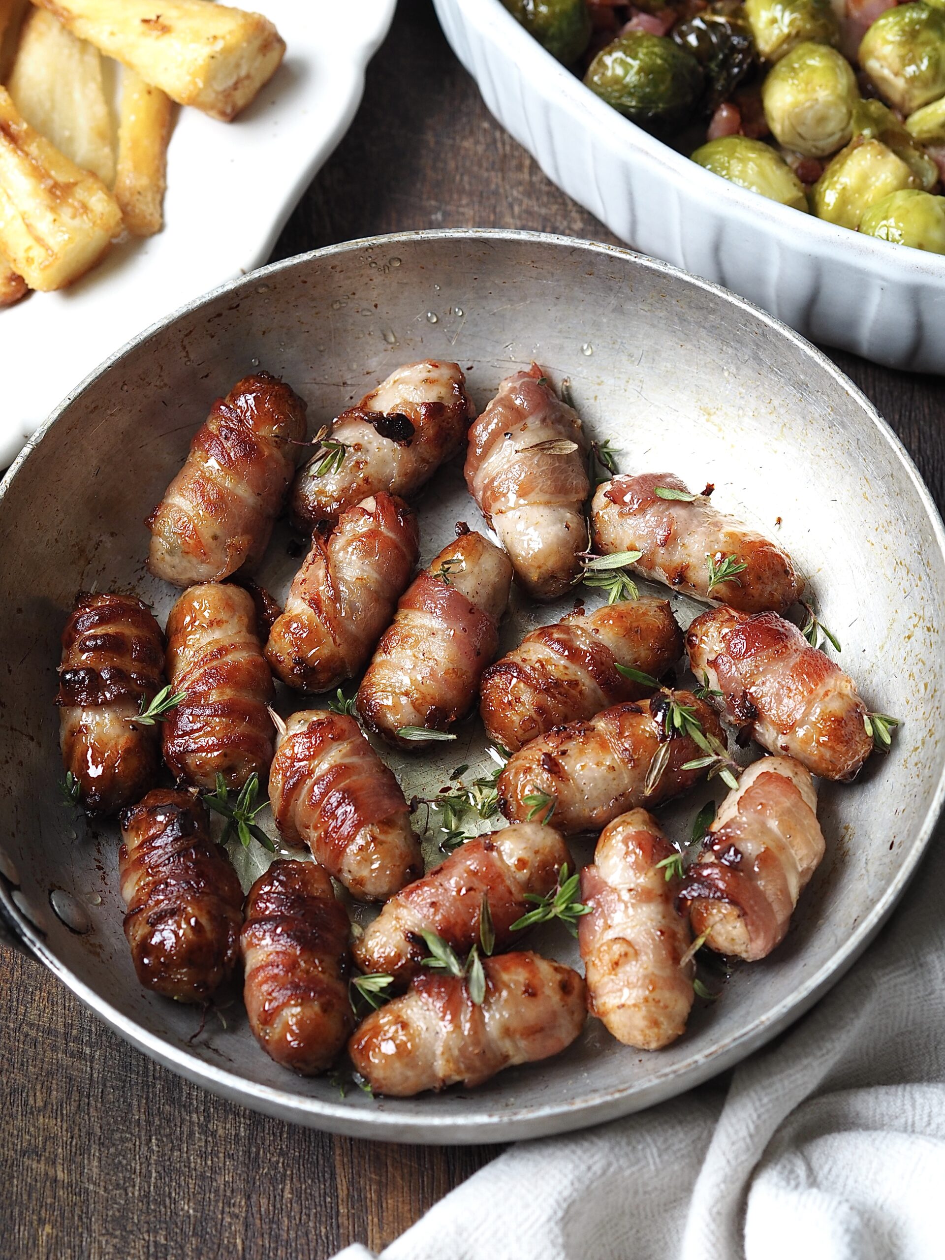 Frying pan of sticky pigs in blankets topped with fresh thyme next to white platters of bacon sprouts and roasted parsnips.