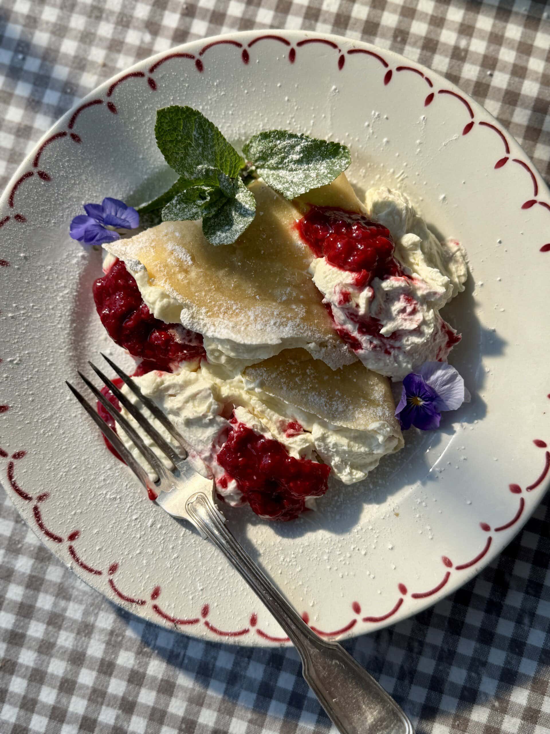 berry crepes on a white plate with raspberries and whipped cheesecake cream.